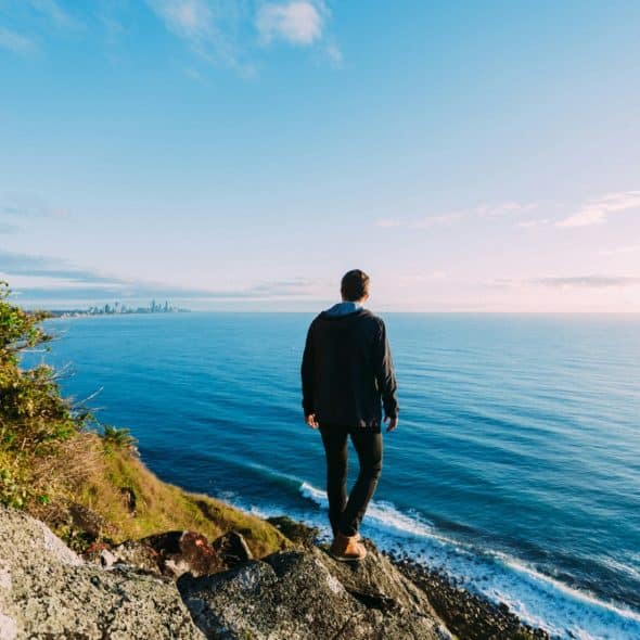 A person standing on a rocky cliff overlooking a vast blue ocean under a bright sky.
