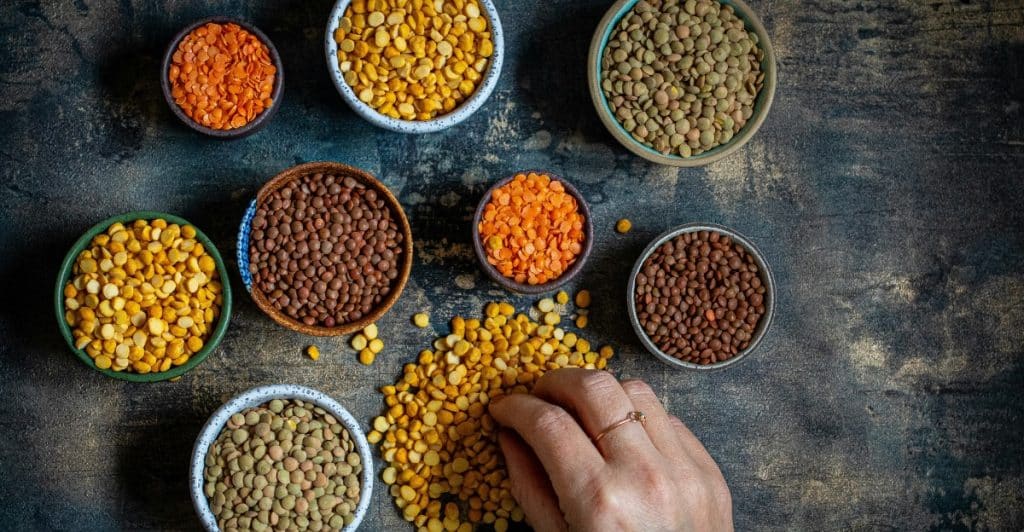 A hand touching a pile of yellow lentils, surrounded by several small bowls filled with different varieties of dried lentils.