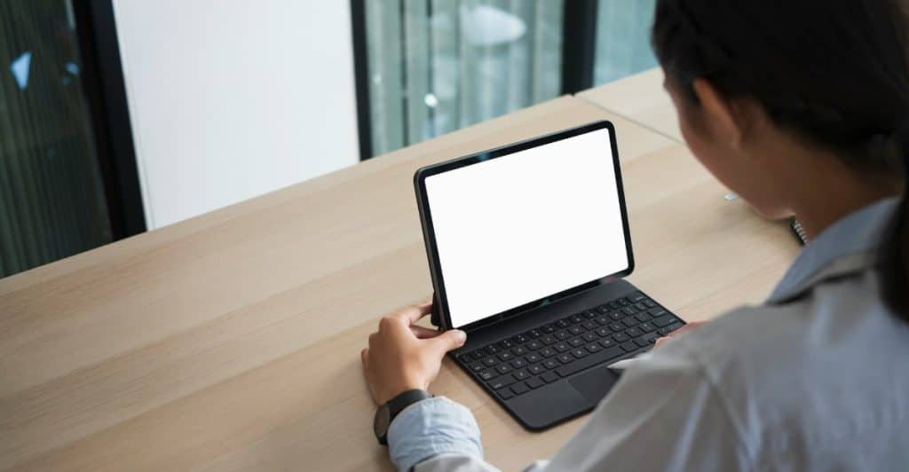 A person seen from over the shoulder, typing on a black Bluetooth keyboard attached to a tablet with a blank screen.