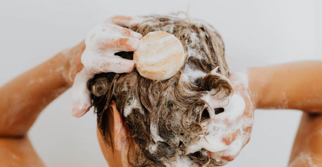 An overhead shot of a person washing their hair with a round, solid shampoo bar, creating a lot of white lather.