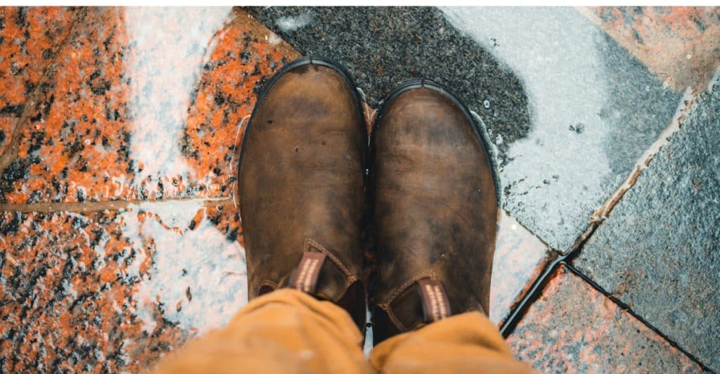 A person's feet are shown from a first-person perspective, wearing brown boots and standing on wet, textured ground.