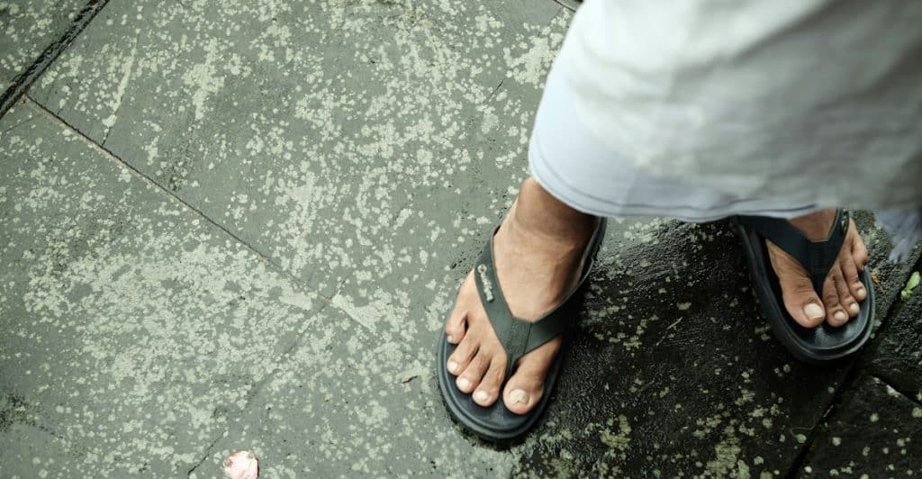 A person's feet are shown from above, wearing black flip-flops on a wet, speckled ground.
