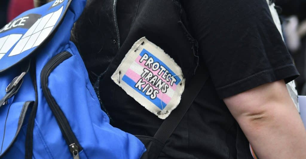 A person wearing a black shirt and a blue backpack with a "PROTECT TRANS KIDS" patch is visible from the chest down.