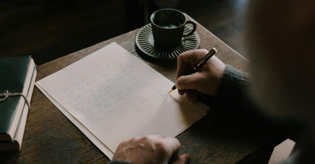 A person's hands writing on a piece of paper with a fountain pen, next to a coffee cup and a journal on a wooden table.