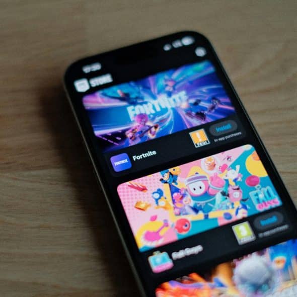 A smartphone showing the Epic Games store with "Fortnite" and "Fall Guys" game listings prominently displayed on a wooden surface.