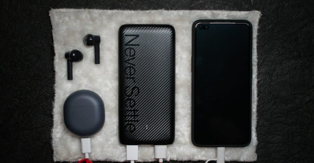 A power bank is charging a smartphone and wireless earbuds, all arranged on a white textured surface.