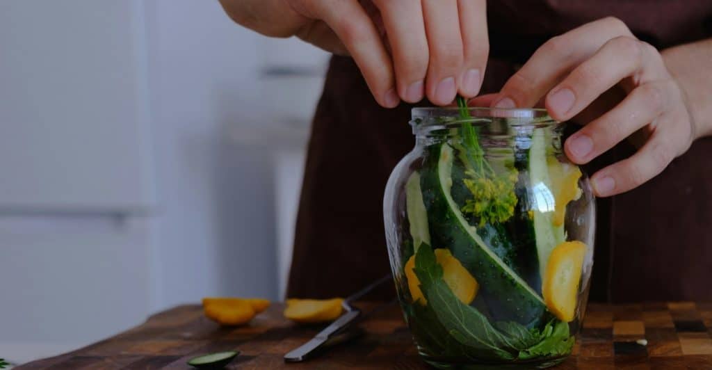 Hands with a wedding ring adding green vegetables and yellow fruit slices to a clear glass jar, preparing a fermentation project on a wooden cutting board.