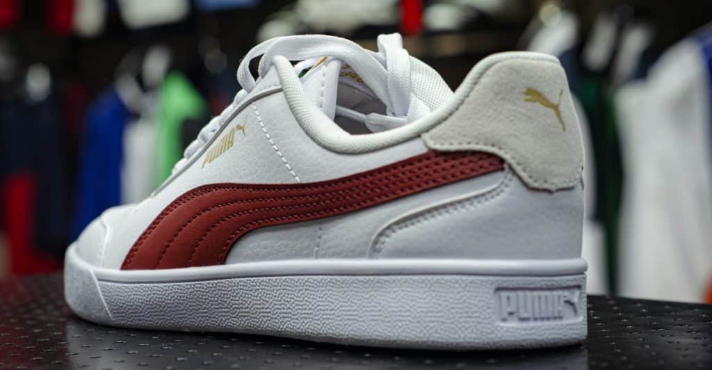 A white and red Puma Suede Classic sneaker with a gold logo on the heel, displayed on a dark surface in front of blurred colorful clothing.