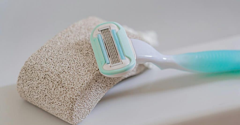 A light blue and white razor leaning against a porous pumice stone on a white surface.
