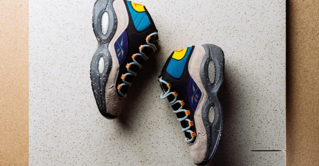 A pair of Reebok Question Mid sneakers with multi-colored accents and a speckled sole, laid out on a light grey textured surface.