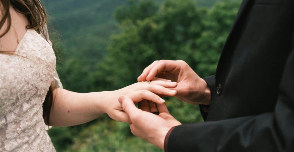 A close-up view of a groom's hands gently placing a wedding ring onto the finger of his bride, who is wearing a white lace wedding dress, during an outdoor ceremony.
