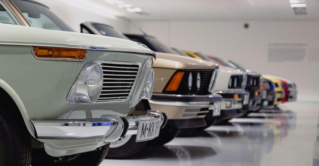 A close-up of the front of several classic cars lined up indoors, showing their headlights and grilles.