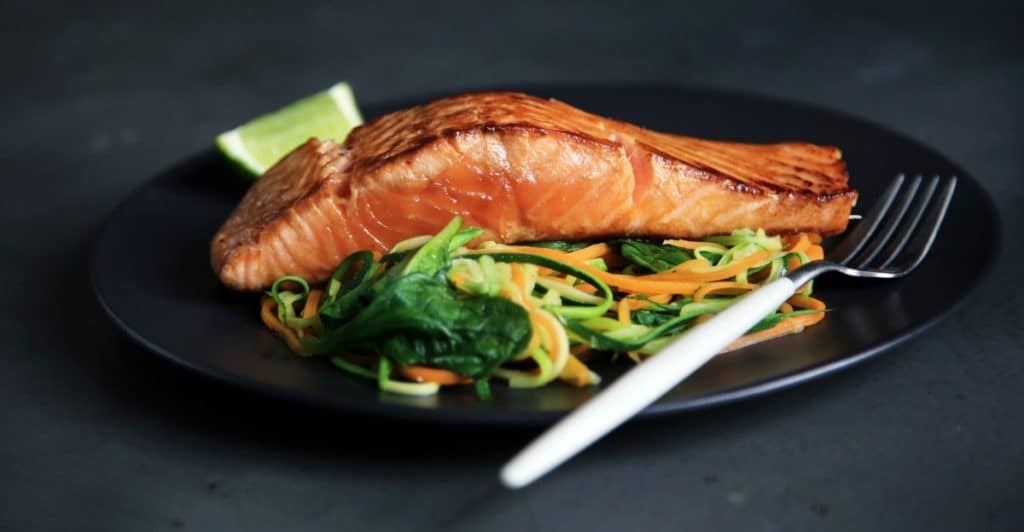 A cooked salmon fillet served on a black plate with a side of mixed spiralized vegetables and a lime wedge.
