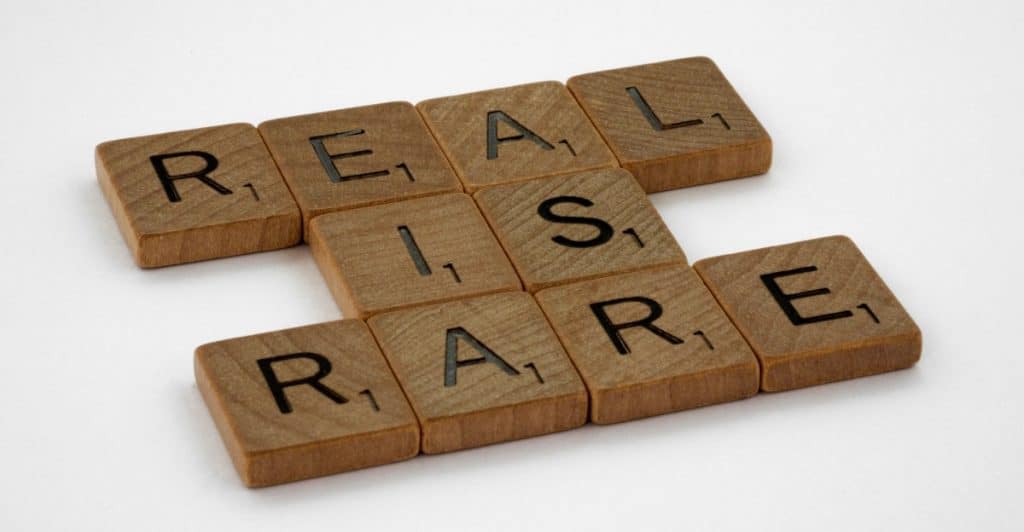 Wooden Scrabble tiles are arranged on a white surface to spell out "REAL IS RARE", with the letters R, E, A, L forming a row and I, S, R, A, R, E intersecting from the 'A' of REAL.