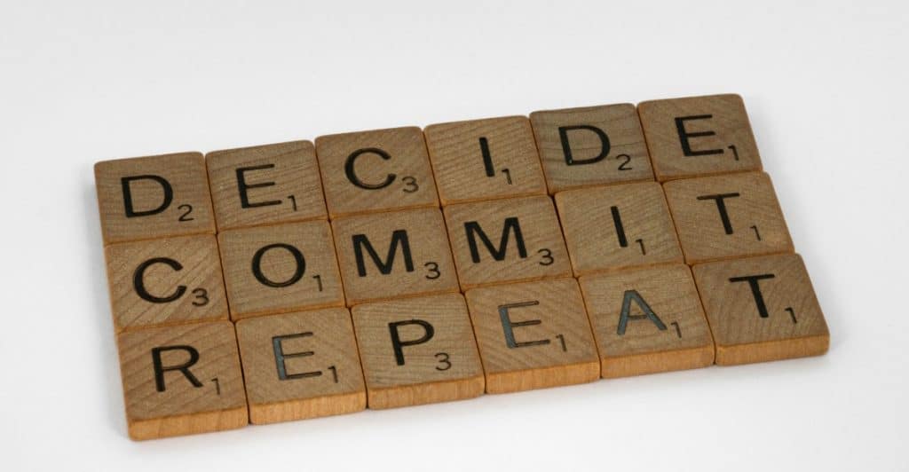 Wooden scrabble tiles are arranged on a white surface to spell out the words "DECIDE", "COMMIT", and "REPEAT".
