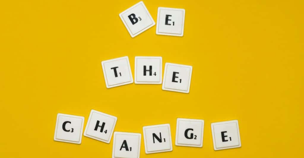 White square tiles with black letters are arranged on a yellow background to spell out the phrase "BE THE CHANGE".