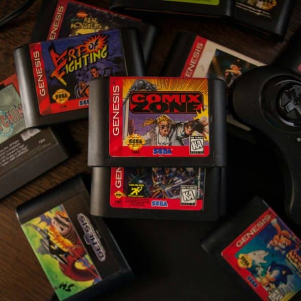 A collection of Sega Genesis video game cartridges including Comix Zone, Art of Fighting, and others, with a black Sega Genesis controller on a dark surface.
