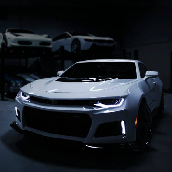 A white sports car with its lights on, parked in a dark garage with other luxury cars on lifts in the background.