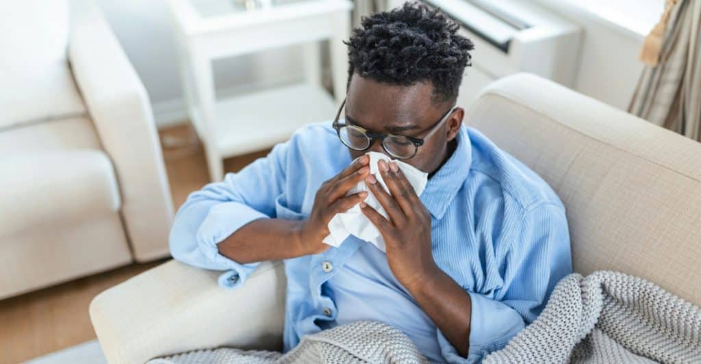 A man who appears to be Black or African American wearing glasses and a blue shirt blowing his nose with a tissue while sitting on a couch with a blanket.
