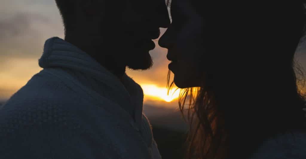 A silhouetted couple embracing with their faces close, illuminated by the setting sun behind them.