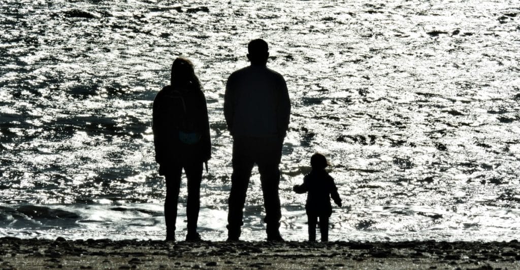 A silhouetted family of three, a man, a woman, and a small child, standing by the water's edge.