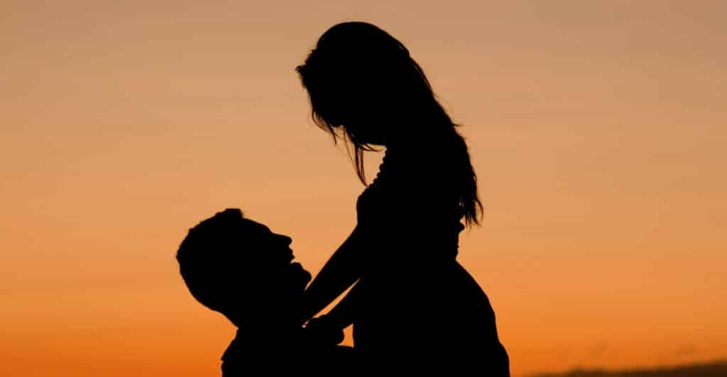 Silhouettes of a man looking up at a woman as he lifts her up, against a sunset sky