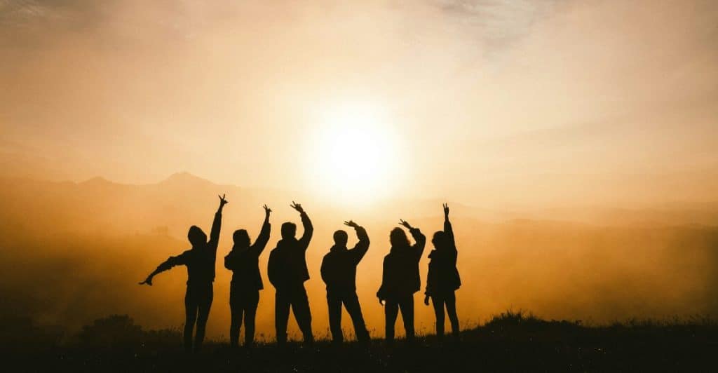 Silhouettes of a group of friends standing on a hill with their arms raised, facing a vibrant sunset.