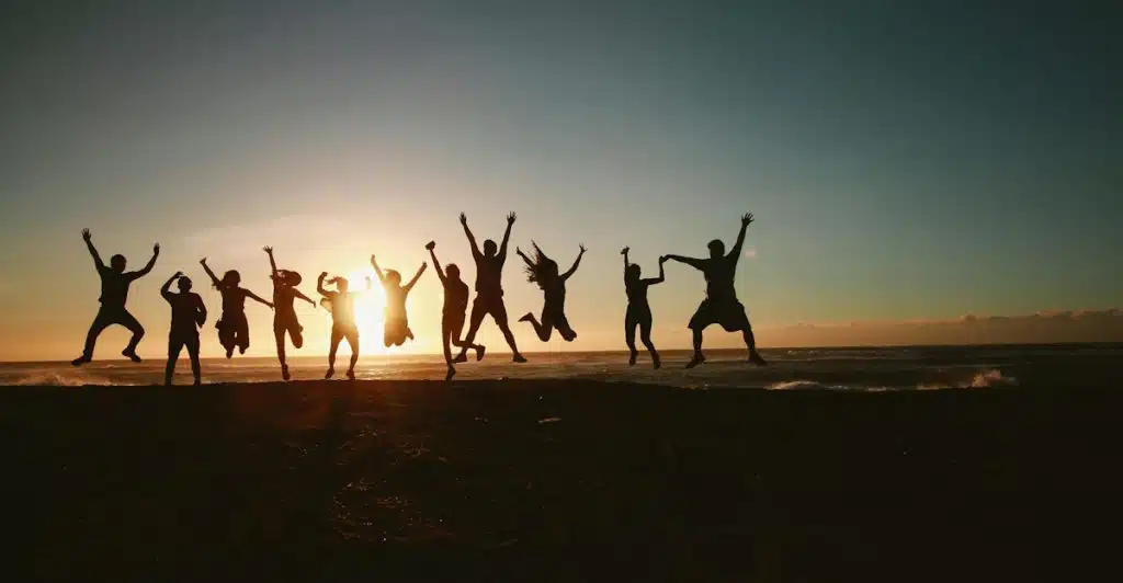 Silhouettes of a group of people jumping joyfully on a beach at sunset, with the sun low on the horizon.