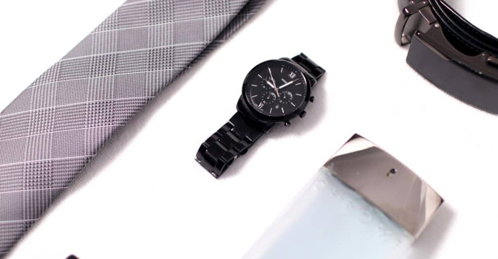 A flat lay of men's accessories including a grey plaid tie, a black watch, a black belt, and a light-colored bottle, on a white surface.