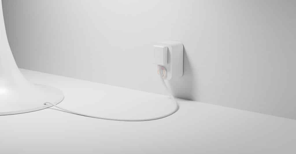 A white smart plug inserted into an outlet on a white wall, with a white cord plugged into it.