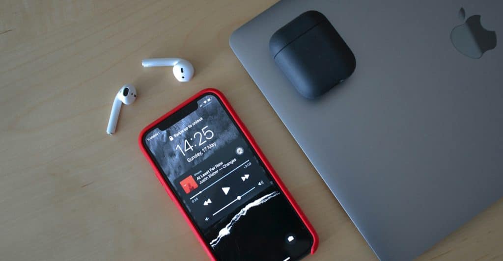 A red-cased smartphone displaying a music player, white AirPods, their charging case, and a silver laptop arranged on a wooden surface.