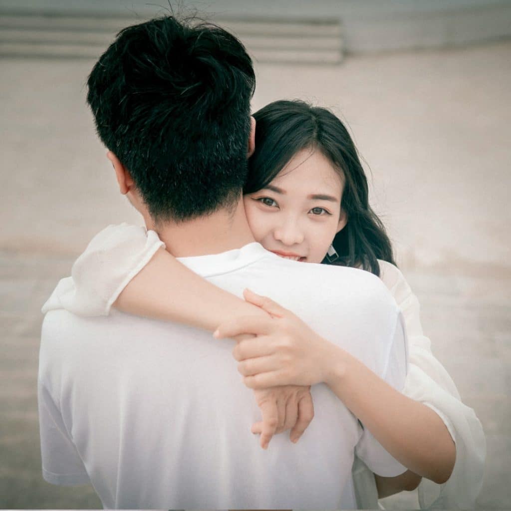 A woman with long dark hair hugging a man , resting her chin on his shoulder and looking at the camera.