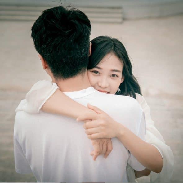 A woman with long dark hair hugging a man , resting her chin on his shoulder and looking at the camera.