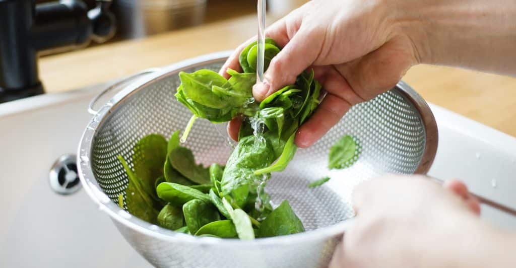 Hands washing fresh spinach leaves in a metal colander under running water in a kitchen sink.