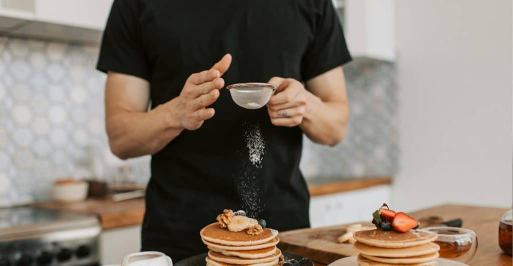 A person's hands holding a sifter, sprinkling powdered sugar over a stack of pancakes topped with berries and nuts.