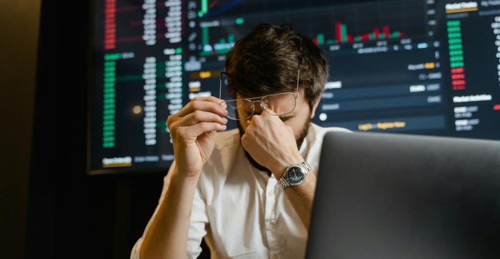 A man with glasses pushed up on his forehead, looking stressed while sitting in front of multiple computer screens displaying data.