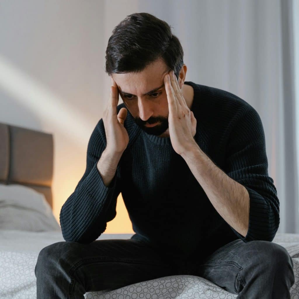 A man sitting on a bed with his hands on his head, looking down with a distressed expression.