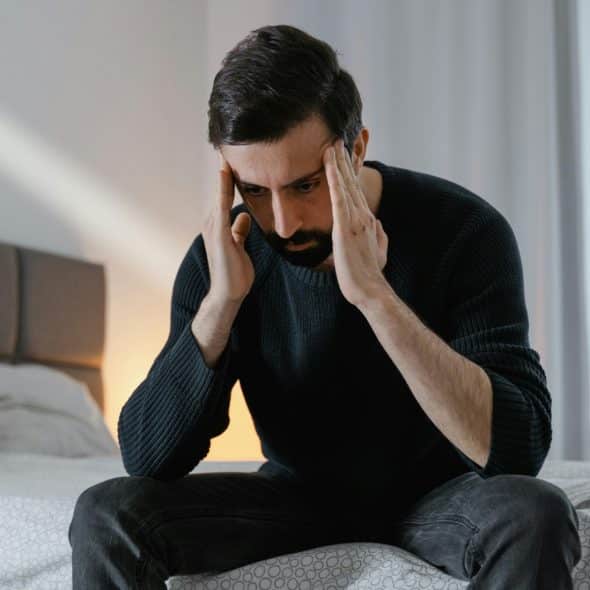 A man sitting on a bed with his hands on his head, looking down with a distressed expression.