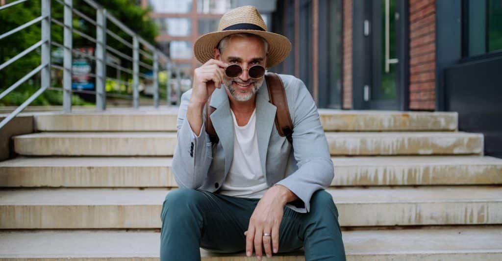 A smiling man with graying hair wearing a hat and sunglasses sitting on outdoor concrete steps.