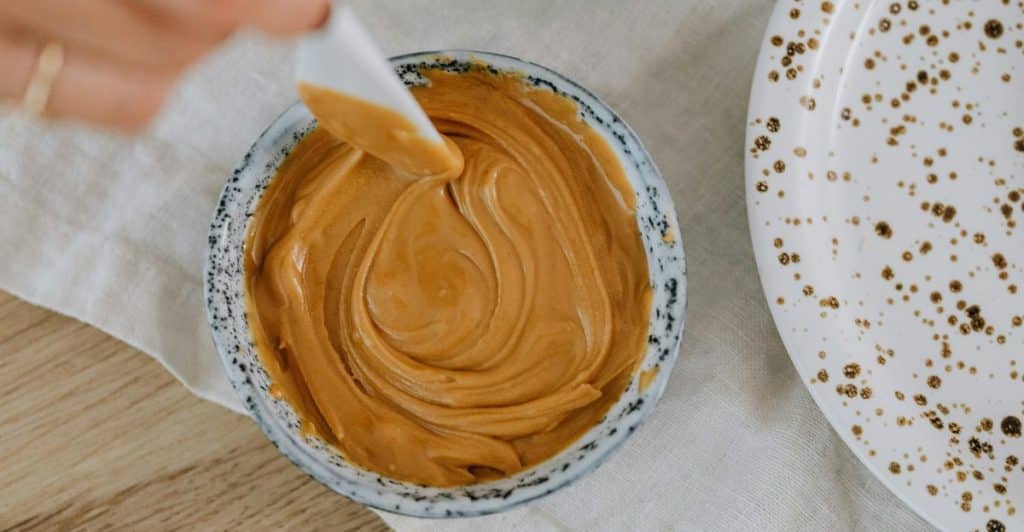 A hand holds a small white spatula, gently swirling creamy peanut butter in a small, patterned bowl, with a speckled white plate and a white linen cloth visible nearby.