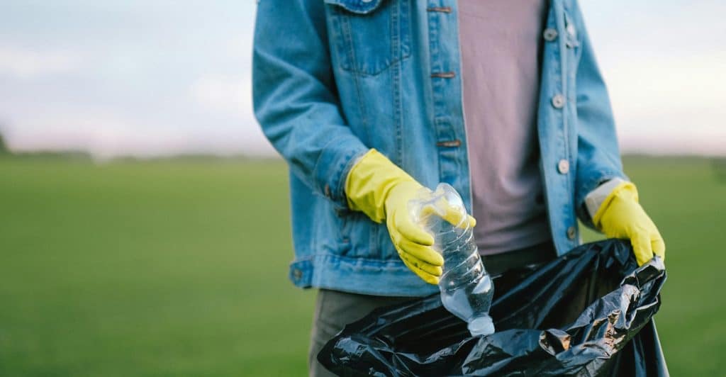 A person wearing a denim jacket and yellow gloves putting a clear plastic bottle into a black trash bag outdoors in a grassy area.