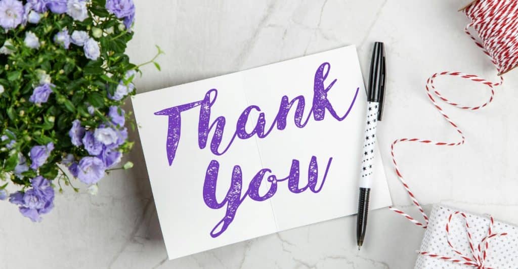 A thank you card with a pen, next to purple flowers and a small gift
