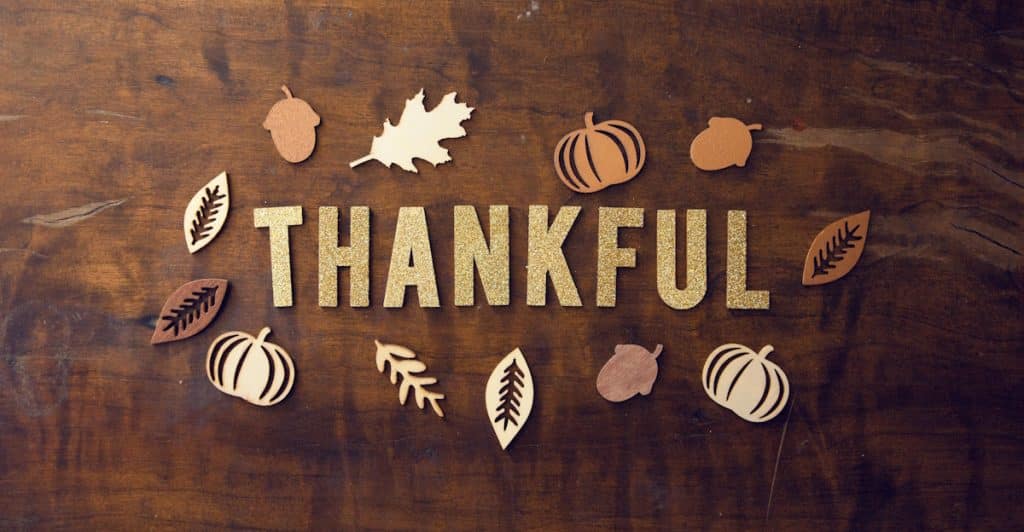 The word "THANKFUL" spelled out in gold, glittery letters on a wooden background, surrounded by decorative wooden cutouts of autumn leaves, acorns, and pumpkins.