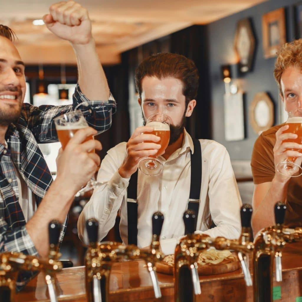 Three men standing at a bar, each holding a glass of beer, with one man raising his fist in celebration while another sips his drink.