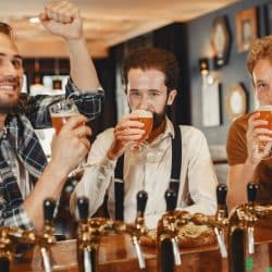 Three men standing at a bar, each holding a glass of beer, with one man raising his fist in celebration while another sips his drink.