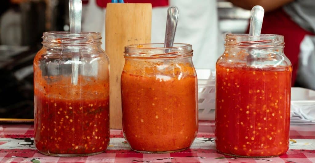 Three glass jars filled with different shades of red and orange homemade hot sauce, each with a spoon, sitting on a checkered tablecloth.