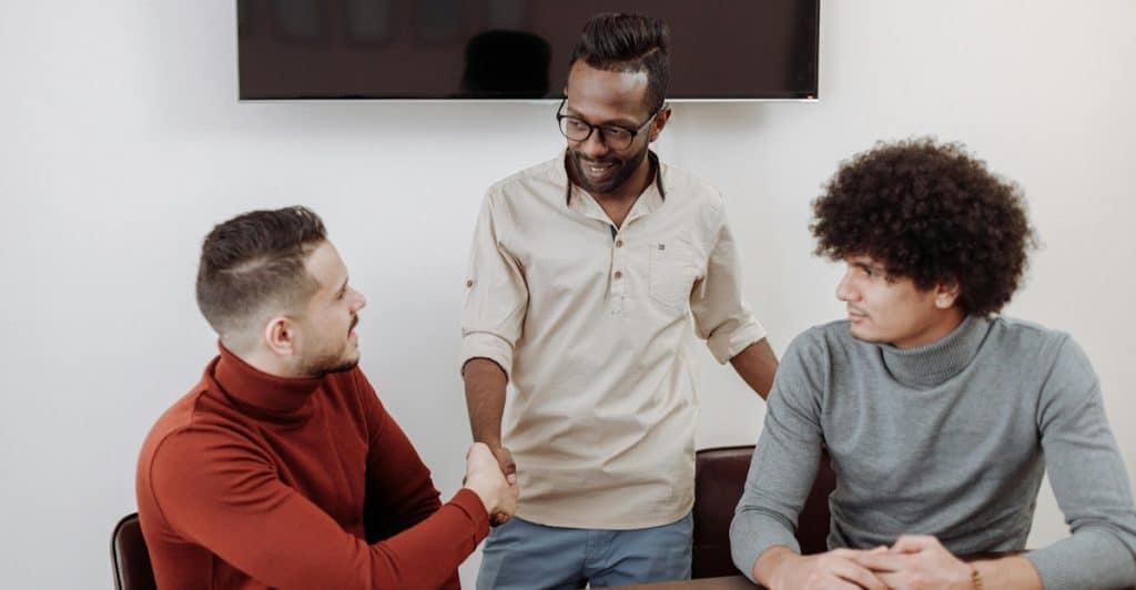In an office setting, two men are shaking hands across a dark wooden table, while a third man with glasses and a beard stands between them, smiling.