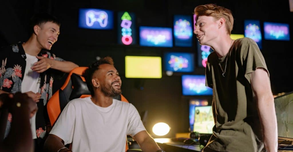 Three young men, two standing and one sitting, laughing and interacting in a dark room filled with glowing screens and gaming equipment.
