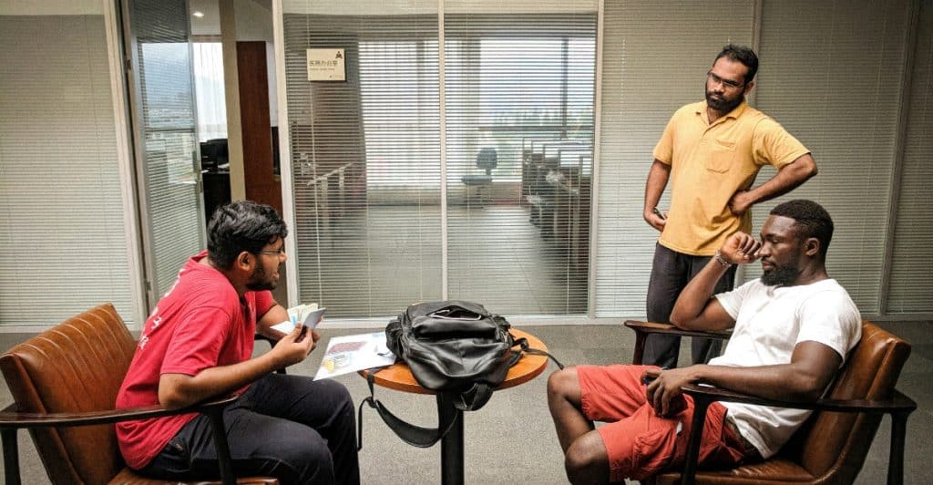Three men in an office setting, with two seated in armchairs talking and one standing and listening.