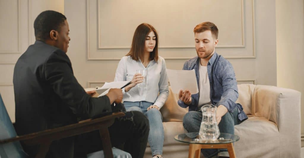Three people are in a room with light walls and a dark gray couch; a man in a dark suit is seated on the left, an unsmiling woman in a white top is in the middle holding a glass of water, and a man in a light blue shirt is on the right, all looking at and discussing documents.